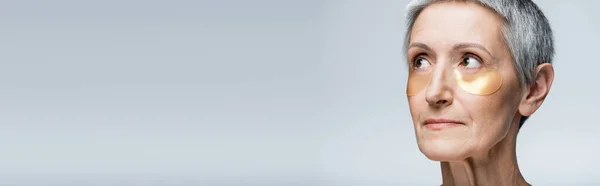 Middle aged woman in golden eye patches looking away isolated on grey, banner — Stock Photo