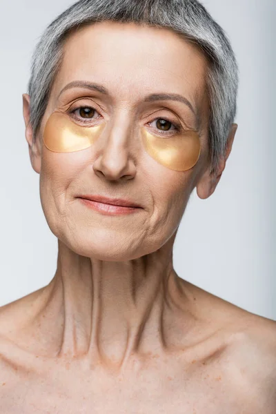 Pleased middle aged woman in golden eye patches looking at camera isolated on grey — Stock Photo