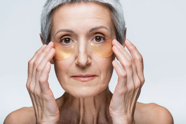 Middle aged woman with bare shoulders applying golden eye patches isolated on grey — Stock Photo