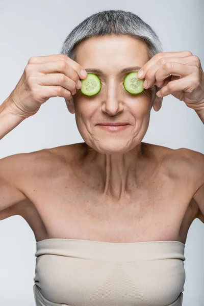 Sonriente mujer de mediana edad cubriendo los ojos con pepinos en rodajas verdes aislados en gris - foto de stock