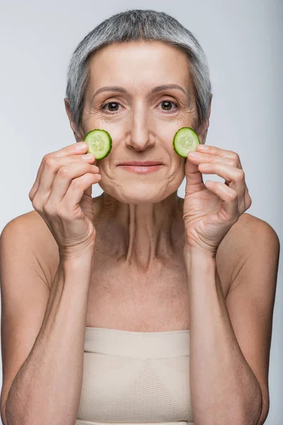 Sonriente mujer de mediana edad sosteniendo pepinos en rodajas verdes aislados en gris - foto de stock