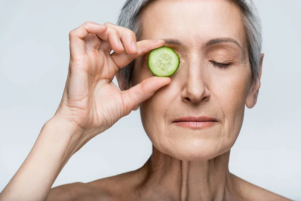 Mujer de mediana edad que cubre el ojo con pepino en rodajas verdes aislado en gris - foto de stock