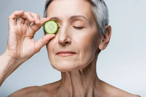 Зрелая женщина закрывает глаза зеленым нарезанным огурцом, изолированным на сером — стоковое фото