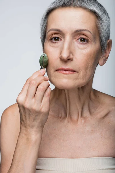 Middle aged woman massaging cheek with jade roller isolated on grey — Stock Photo