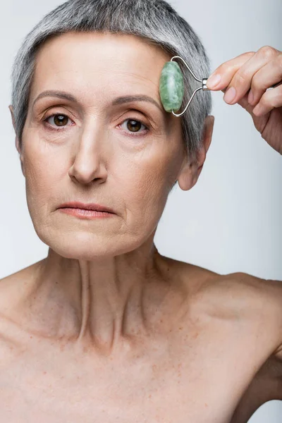 Mujer de mediana edad masajeando la frente con rodillo de jade aislado en gris - foto de stock