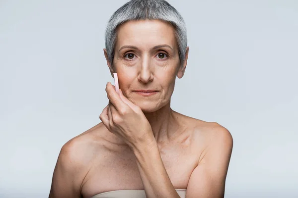 Middle aged woman with bare shoulders applying makeup foundation with beauty sponge isolated on grey — Stock Photo