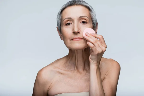 Жінка середнього віку з сірим волоссям наносить макіяж фундамент з красивою губкою ізольовані на сірому — стокове фото