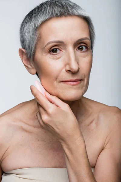 Donna di mezza età con capelli grigi in possesso di spugna di bellezza mentre si applica fondotinta isolato su grigio — Foto stock