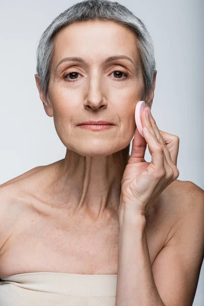 Mature woman with grey hair holding beauty sponge and applying face foundation isolated on grey — Stock Photo