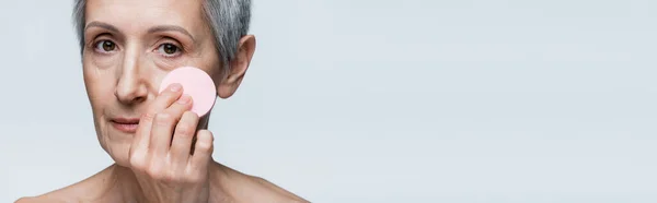 Primo piano di donna matura con capelli grigi in possesso di spugna di bellezza e l'applicazione di fondazione del viso isolato su grigio, banner — Foto stock