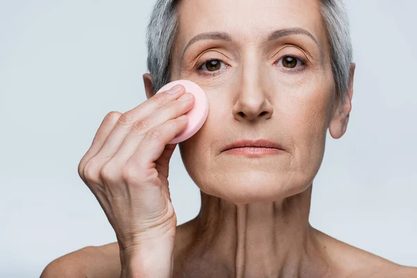 Mujer madura sosteniendo esponja de belleza y aplicando maquillaje fundación aislado en gris - foto de stock