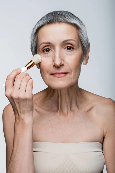 Mature woman with grey hair applying face powder with cosmetic brush isolated on grey — Stock Photo