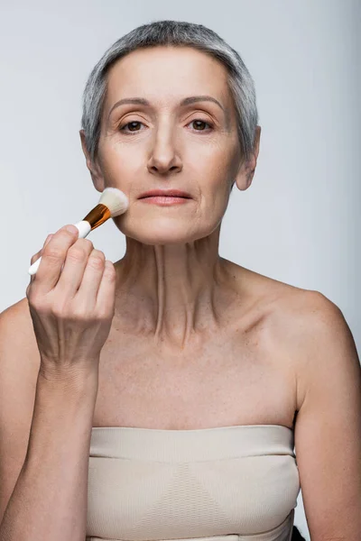 Middle aged woman with grey hair applying face powder with cosmetic brush isolated on grey — Stock Photo
