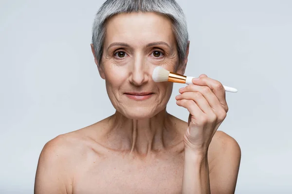 Smiling mature woman with grey hair applying face powder with cosmetic brush isolated on grey — Stock Photo