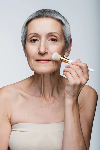 Pleased mature woman with grey hair applying face powder with cosmetic brush isolated on grey — Stock Photo