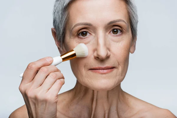 Mature woman with bare shoulders applying face powder on cheek with cosmetic brush isolated on grey — Stock Photo
