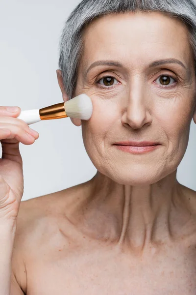 Mujer de mediana edad con hombros desnudos aplicando polvo facial en la mejilla con cepillo cosmético aislado en gris - foto de stock