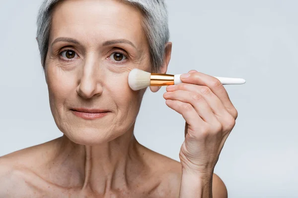 Mulher de meia idade aplicando pó facial isolado no cinza — Fotografia de Stock