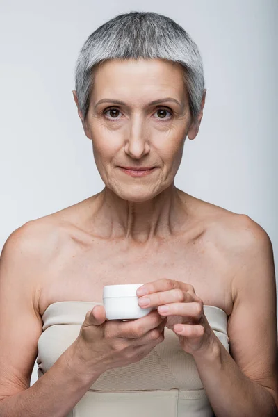 Mulher madura com ombros nus segurando recipiente com creme facial isolado em cinza — Fotografia de Stock