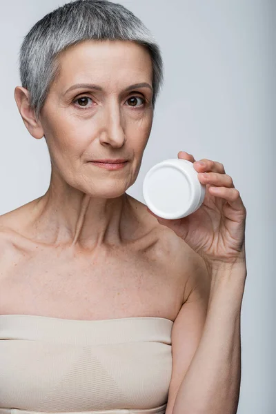 Mulher de meia idade com ombros nus segurando recipiente com creme facial isolado em cinza — Fotografia de Stock