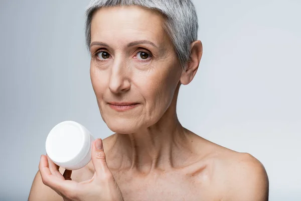 Mujer madura con pelo gris sosteniendo contenedor con crema facial aislado en gris - foto de stock