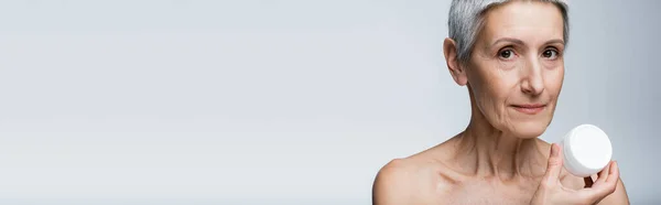 Mature woman with grey hair holding container with face cream isolated on grey, banner — Stock Photo
