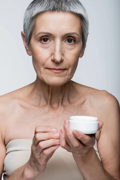 Femme mûre avec récipient de tenue de cheveux gris avec crème cosmétique isolé sur gris — Photo de stock