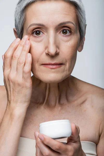 Mature woman with grey hair applying anti-wrinkle cream isolated on grey — Stock Photo