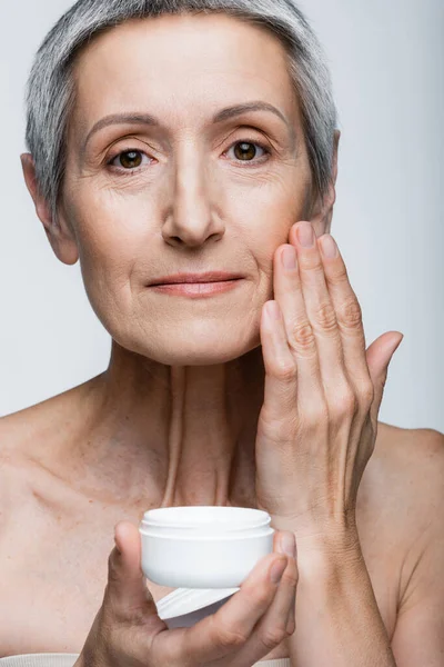 Mulher de meia idade com cabelos grisalhos aplicando creme anti-rugas isolado no cinza — Fotografia de Stock