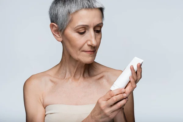 Femme d'âge moyen avec les cheveux gris regardant tube avec lotion isolé sur gris — Photo de stock
