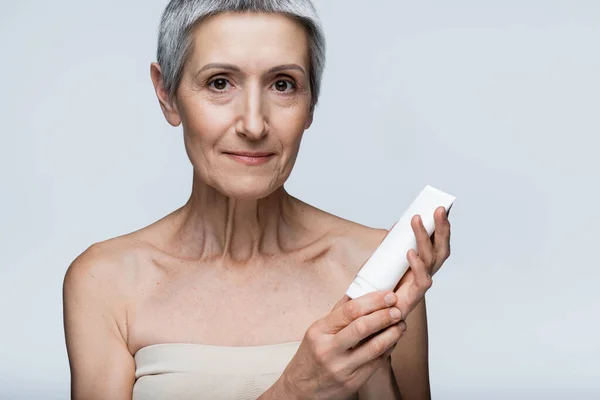 Mature woman with grey hair holding tube with lotion isolated on grey — Stock Photo