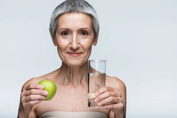 Cheerful middle aged woman holding glass of water and green apple isolated on grey — Stock Photo