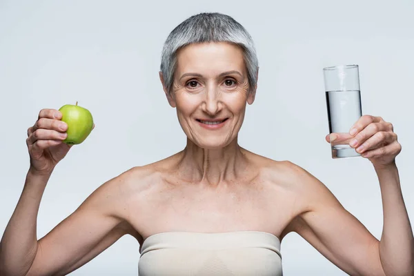 Happy middle aged woman holding glass of water and green apple isolated on grey — Stock Photo