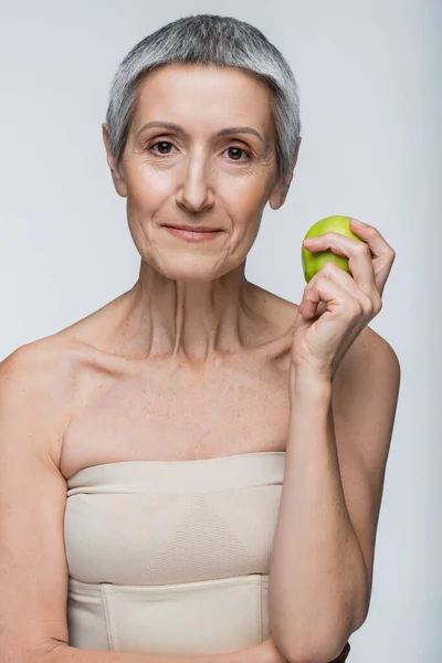 Sonriente mujer de mediana edad sosteniendo manzana verde aislada en gris - foto de stock