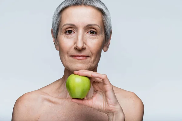 Middle aged woman with bare shoulders holding green apple isolated on grey — Stock Photo