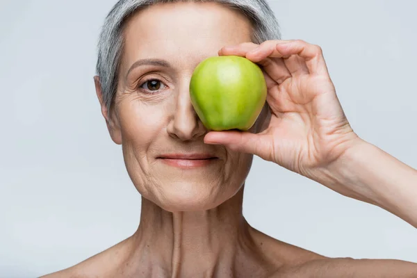 Smiling middle aged woman covering eye with green apple isolated on grey — Stock Photo