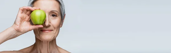 Lächelnde Frau mittleren Alters, die Augen mit grünem Apfel auf grauem Hintergrund bedeckt, Banner — Stockfoto