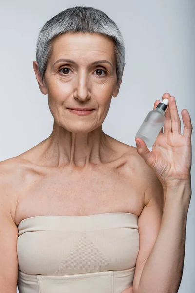 Middle aged woman with grey hair holding bottle with serum isolated on grey — Stock Photo