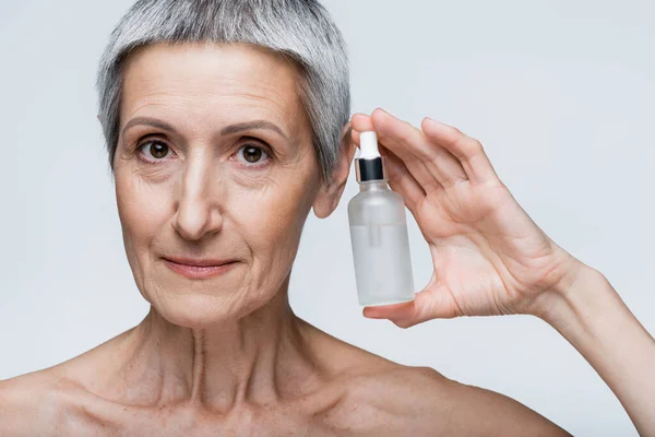 Mature woman with grey hair holding bottle with serum isolated on grey — Stock Photo