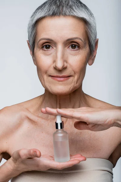 Middle aged woman with grey hair holding bottle with moisturizing serum isolated on grey — Stock Photo