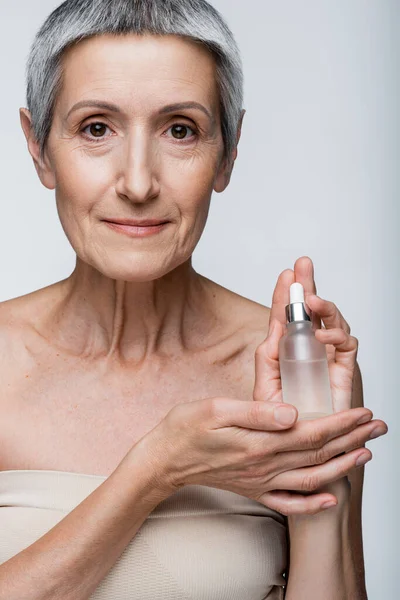 Heureux femme mature avec cheveux gris tenant bouteille avec sérum hydratant isolé sur gris — Photo de stock