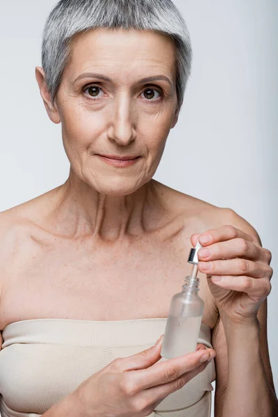 Femme mature aux cheveux gris tenant bouteille et pipette avec sérum hydratant isolé sur gris — Photo de stock