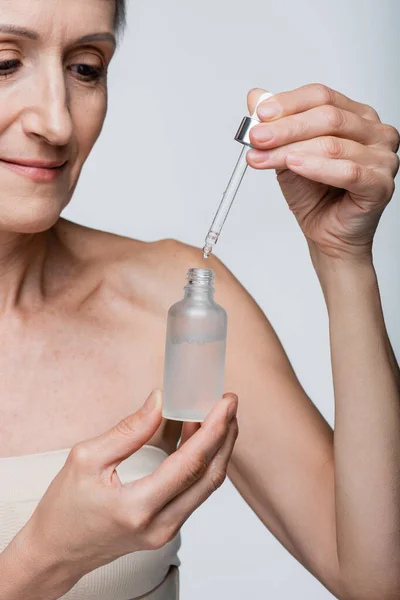 Mujer alegre y madura sosteniendo botella y pipeta con suero hidratante aislado en gris - foto de stock