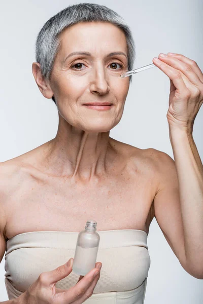 Sorridente donna di mezza età che tiene la bottiglia e applica il siero isolato sul grigio — Foto stock