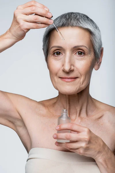 Веселая женщина средних лет с седыми волосами, держа бутылку и применяя сыворотку, изолированную на сером — стоковое фото