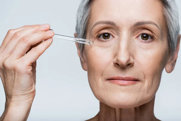 Nahaufnahme einer Frau mittleren Alters mit grauem Haar, die feuchtigkeitsspendendes Serum auf grau isoliert aufträgt — Stockfoto