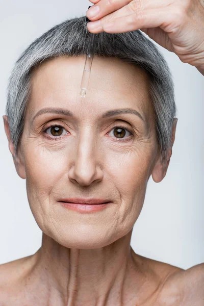 Vista da vicino della donna di mezza età che applica siero di vitamina C isolato sul grigio — Foto stock