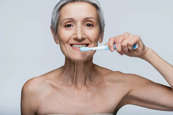 Весела жінка середнього віку тримає зубну щітку і чистить зуби ізольовано на сірому — стокове фото