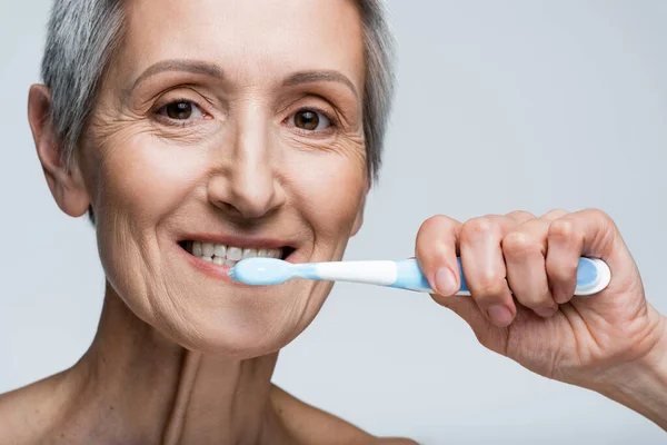 Smiling middle aged woman holding toothbrush and brushing teeth isolated on grey — Stock Photo