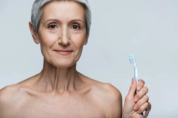 Cheerful middle aged woman holding toothbrush isolated on grey — Stock Photo